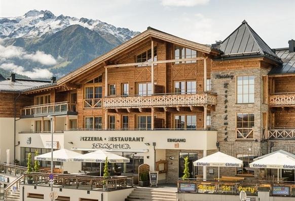 Ein traditionelles Bergrestaurant mit einer großen Terrasse und Sonnenschirmen. Im Hintergrund sind majestätische Berge und ein klarer Himmel zu sehen.
