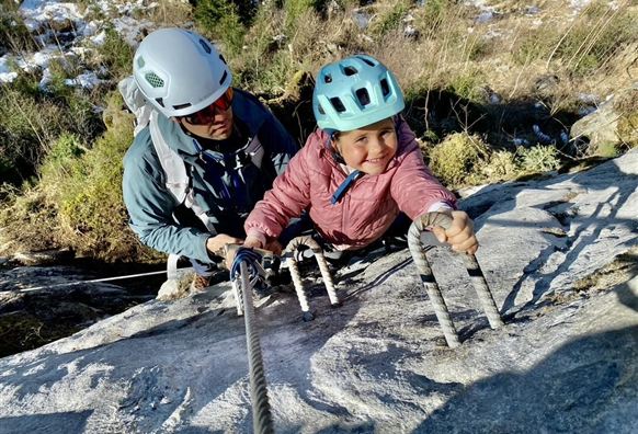 A climber secures another climber at a rocky wall. The scene shows a clear blue sky and an impressive mountain landscape.