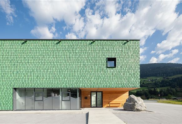 Ein modernes Gebäude mit einer grünen Schindelfassade und großen Fenstern. Im Hintergrund sieht man eine hügelige Landschaft unter einem klaren Himmel.