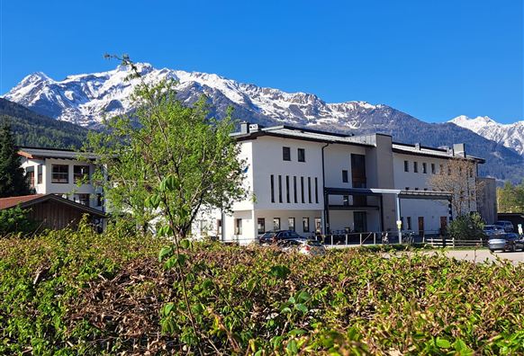 Ein modernes Gebäude vor einer beeindruckenden Bergkulisse. Die Landschaft ist grün und sonnig, mit schneebedeckten Gipfeln im Hintergrund.