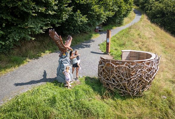 Ein Weg führt durch eine grüne Landschaft. Zwei Kinder stehen neben einer großen Statue eines Adlers und einer Strukturen aus Zweigen.