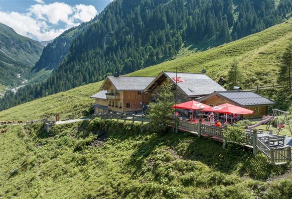 Eine alpenländische Hütte in einer grünen Berglandschaft. Entlang der Hütte stehen rote Sonnenschirme und der Himmel ist klar und blau.