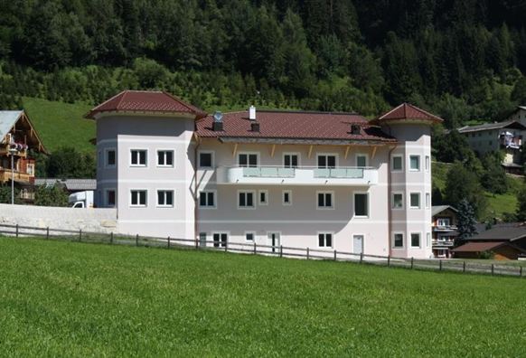 Ein modernes Gebäude umgeben von grünen Wiesen und Bergen. Das Haus hat eine helle Fassade und einen roten Dachstuhl.