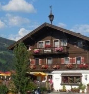Een charmant, twee verdiepingen tellend houten huis met balkons en kleurrijke bloembakken. Op de achtergrond zijn idylische bergen en een blauwe lucht te zien.