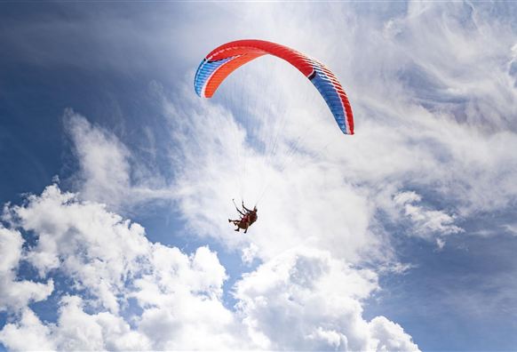 A paraglider floats through the blue sky with white, fluffy elements. The parachute has a red-blue design.