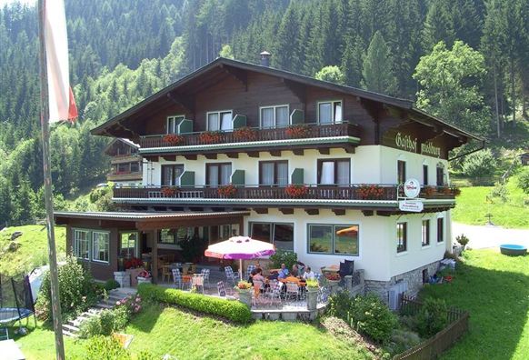 Ein malerisches Gästehaus in den Bergen, umgeben von grüner Natur. Auf der Terrasse sind Tische und Stühle mit Sonnenschirmen.