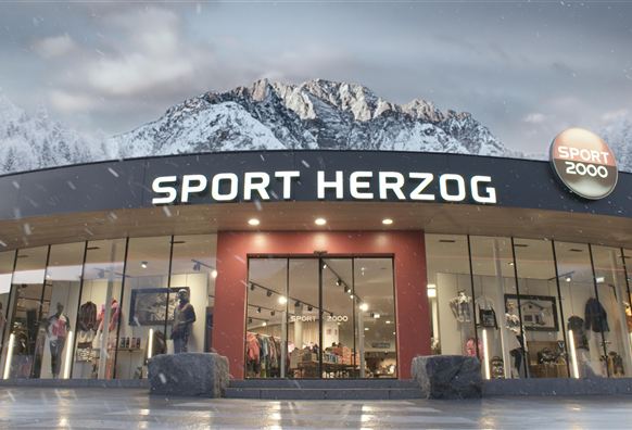 Ein sportliches Geschäft namens "Sport Herzog", umgeben von schneebedeckten Bergen. Die Fassade ist modern und einladend.