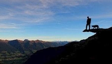 Eine Person steht mit einem Hund auf einem Felsen und blickt in die weite Landschaft. Der Himmel ist klar und die Berge sind im Hintergrund sichtbar.