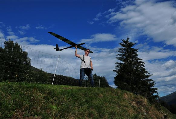 Een man staat op een heuvel en houdt een modelvliegtuig in zijn hand. Op de achtergrond zijn een blauwe lucht en enkele bomen te zien.