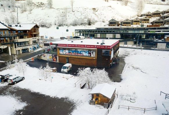 Ein schneebedecktes Dorf mit einem modernen Gebäude und mehreren Skihütten. Autos stehen auf dem Parkplatz, umgeben von einer winterlichen Landschaft.