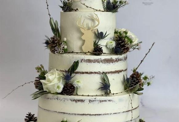 Eine elegante Torte mit mehreren Schichten, dekoriert mit Blumen und Tannenzapfen. Oben befindet sich ein Hirsch-Design aus Holz.