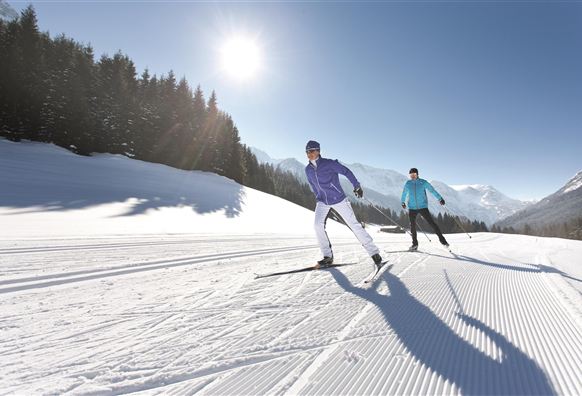 Twee snowboarders rijden over een witte piste in de zon. Op de achtergrond zijn besneeuwde bergen en bomen te zien.