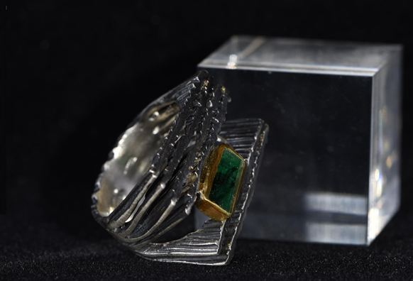 An elegant silver ring with a green stone and golden accents. The ring stands next to a clear, square block.