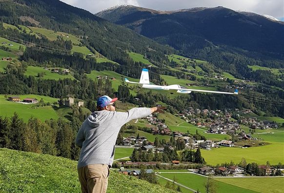 Een man staat op een weiland en wijst naar een stad in de vallei. De omgeving is groen met bergen op de achtergrond en een blauwe lucht.