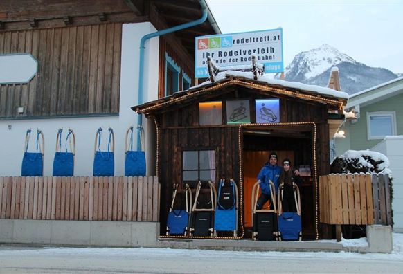 A wooden building with an entrance in front of which two people are standing. Next to the entrance, sleds and blue backpacks are displayed, while snow-covered mountains can be seen in the background.