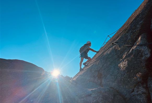 Ein Kletterer erklimmt einen steilen Felsen unter einem klaren blauen Himmel. Die Sonne strahlt im Hintergrund und erzeugt eine schöne Lichtstimmung.