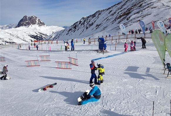 Eine verschneite Berglandschaft mit vielen Skifahrern und snowboardenden Kindern. Im Hintergrund sind hohe Berge und ein klarer Himmel zu sehen.