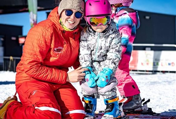 Eine lächelnde Skilehrerin in einem roten Anzug hilft einem kleinen Mädchen beim Skifahren. Im Hintergrund stehen weitere Kinder auf einer schneebedeckten Piste.