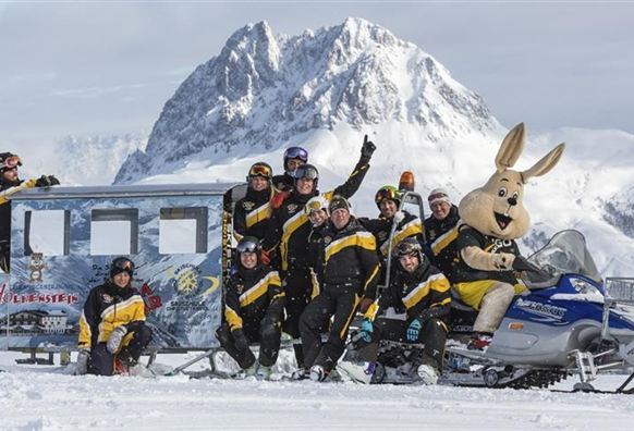 Eine Gruppe von Menschen in winterlicher Kleidung posiert vor einem Schneemobil in einer schneebedeckten Landschaft. Im Hintergrund sieht man majestätische Berge.