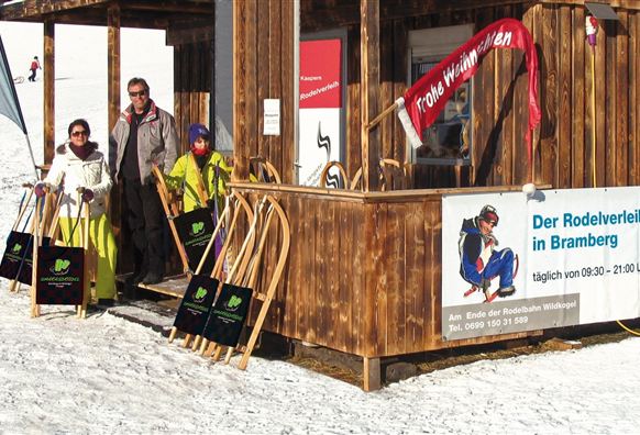 A wooden stand with a sled rental in the mountains. Family members are standing in the snow with sleds in hand.