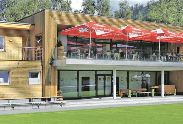 Ein modernes Holzgebäude mit einer großen Terrasse und roten Markisen. Die Umgebung ist grün und bietet eine schöne Aussicht.