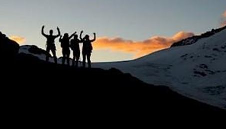 Eine Gruppe von Wanderern steht silhouettiert gegen den Sonnenuntergang. Hinter ihnen sind Berge und Schnee zu sehen.