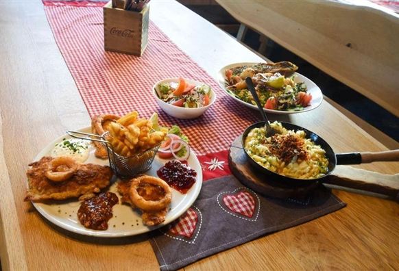 Ein Festmahl mit verschiedenen Gerichten auf einem Tisch. Es sind Pommes, Salat, ein Hauptgericht mit Fleisch und Sauce sowie eine kleine Pfanne mit Beilage zu sehen.