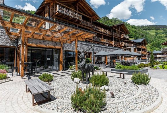 Eine moderne Lodge mit Holzfassade und großen Fenstern umgeben von Grünanlagen. Im Vordergrund befindet sich ein dekorativer Garten mit einem Steinbecken und Sitzgelegenheiten.