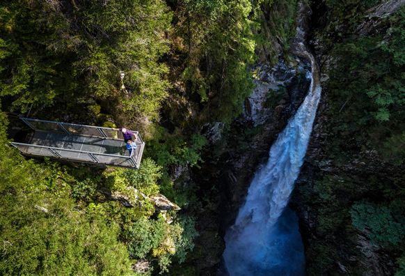 A majestic waterfall flows through a green gorge. Surrounded by dense trees and lush vegetation, the water cascades down.
