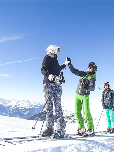 A group of skiers stands on a snowy mountain in the sun. In the background, there are snow-covered mountains and a clear blue sky.