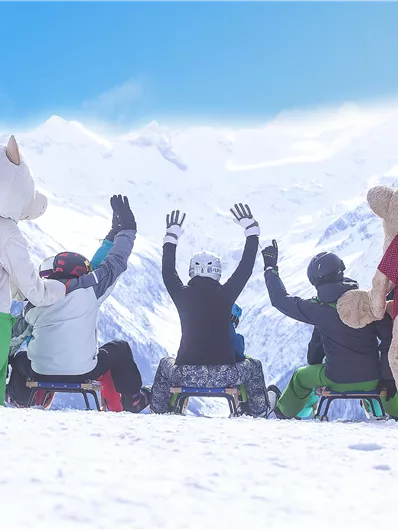 Eine Gruppe von Menschen in bunten Wintersportanzügen sitzt auf Schlitten im Schnee. Im Hintergrund sind schneebedeckte Berge und ein blauer Himmel zu sehen.