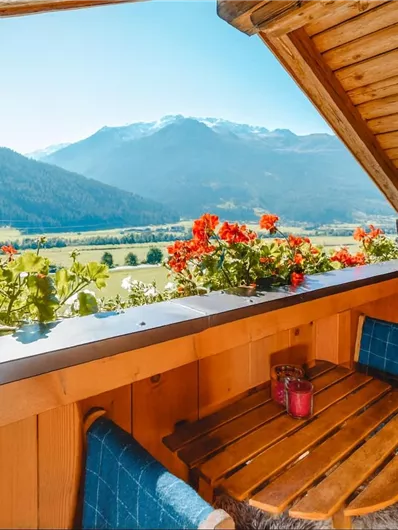 A cozy balcony with wooden cladding and blooming flowers. The view extends over a picturesque valley with mountains in the background.