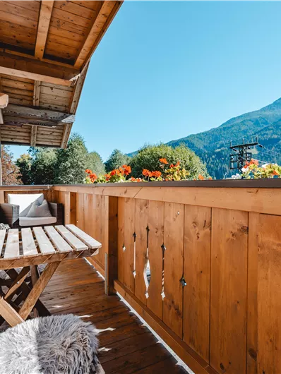 A cozy balcony with a wooden railing and flowers. In the background, mountains and a clear blue sky can be seen.