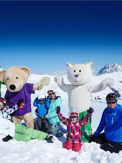 A group of people is standing in the snow, surrounded by mountains. Two bear costumes are posing with the children and adults, while everyone happily waves at the camera.