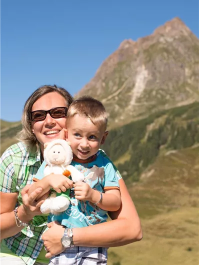 A woman and a boy are standing in front of a mountainous landscape. The boy is holding a stuffed animal in his arms and both are smiling happily.