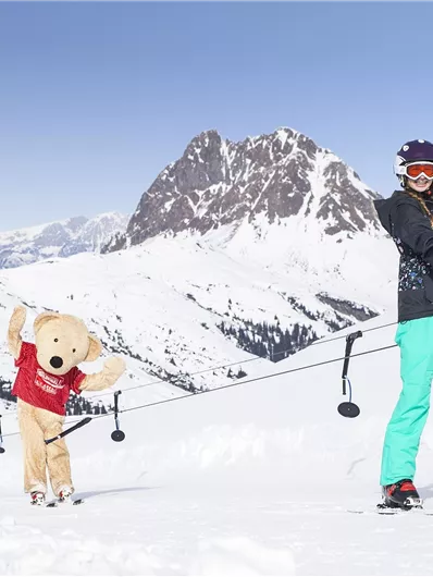 Eine Skifahrerin mit Sonnenbrille und bunten Skihosen winkt fröhlich. Neben ihr steht ein großer, brauner Stoffbär in einem roten T-Shirt im Schnee.
