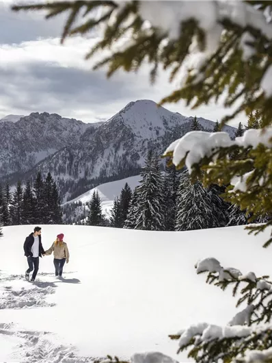 Ein Paar spaziert durch eine verschneite Landschaft mit Bergen im Hintergrund. Der Himmel ist teilweise bewölkt und umgeben von grünen Nadelbäumen.