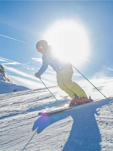 Ein Skifahrer fährt durch den Schnee unter einem strahlend blauen Himmel. Die Sonne scheint hell hinter ihm.