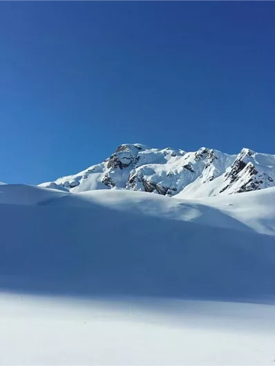 Een met sneeuw bedekt landschap onder een heldere blauwe lucht. Op de achtergrond torenen majestueuze bergen eruit.