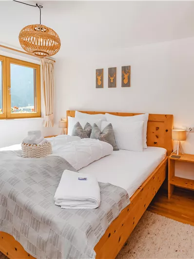 A cozy bedroom with a large wooden bed and light curtains. The room has windows that provide plenty of light and a view of the mountains.