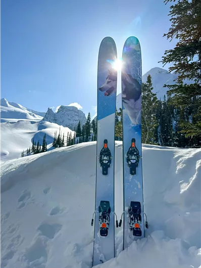 Two skis are standing in the snow, with mountains in the background. The sun is shining brightly and the landscape is wintry and peaceful.