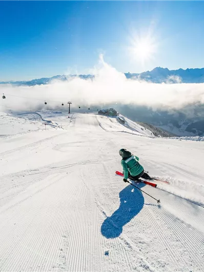 Ein Skifahrer fährt eine perfekt präparierte Piste hinunter. Im Hintergrund sind Berge und ein strahlend blauer Himmel zu sehen.