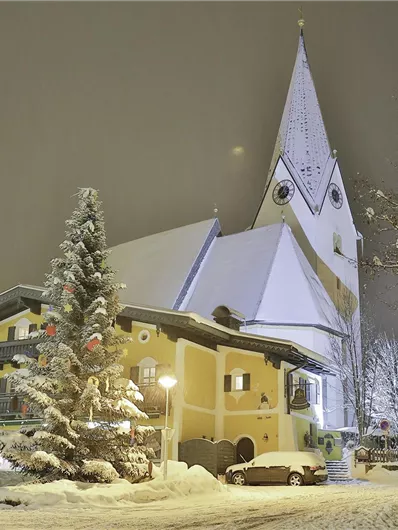 A snowy winter landscape with a charming yellow building and a church in the background. The sky is overcast and the surroundings are festively illuminated.