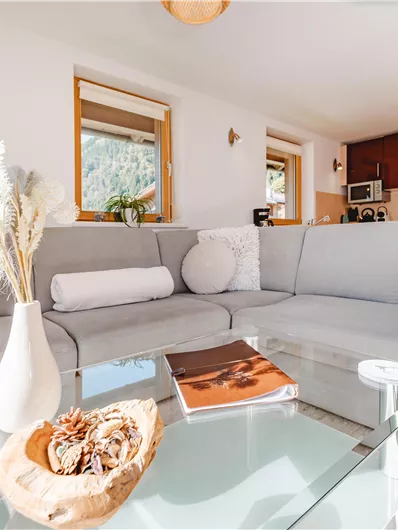A modern living room with a gray couch and a glass table. In the background, there is a kitchen and windows letting in daylight.