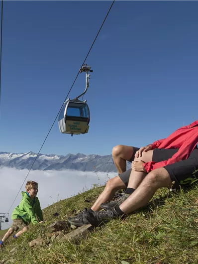 Eine Familie entspannt auf einer grünen Wiese in den Bergen. Eine Seilbahn schwebt über den Nebel im Tal.