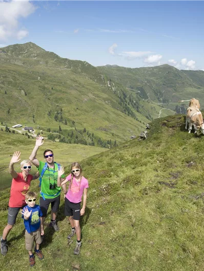 Eine Familie steht fröhlich auf einer grünen Wiese in den Bergen. Im Hintergrund sind sanfte Hügel und Kühe zu sehen.