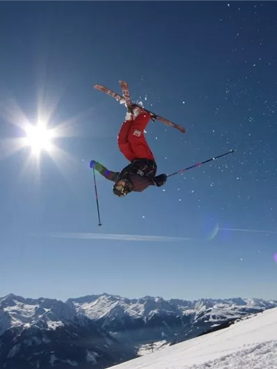 Ein Skifahrer führt einen Sprung in der Luft aus. Der Himmel ist klar und die schneebedeckten Berge sind im Hintergrund sichtbar.