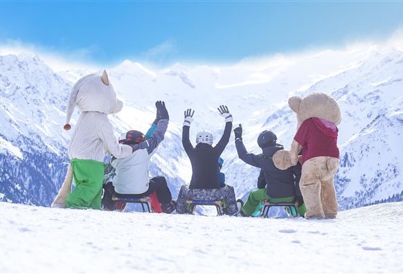 Eine Gruppe von Menschen in bunten Wintersportanzügen sitzt auf Schlitten im Schnee. Im Hintergrund sind schneebedeckte Berge und ein blauer Himmel zu sehen.