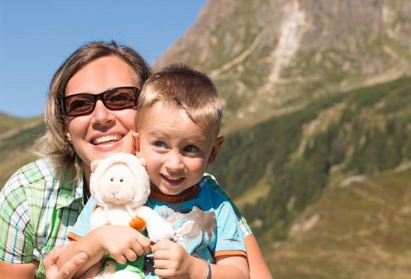 A woman and a boy are standing in front of a mountainous landscape. The boy is holding a stuffed animal in his arms and both are smiling happily.