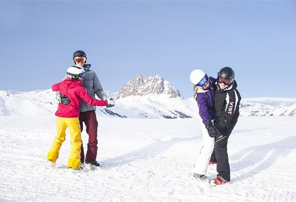 Een groep van vier personen staat op een besneeuwde piste. De zon schijnt en op de achtergrond zijn besneeuwde bergen te zien.